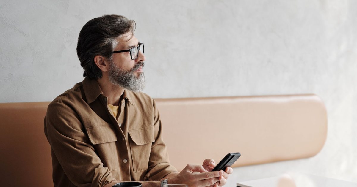 Bearded man in a café holding a phone, looking away in thought