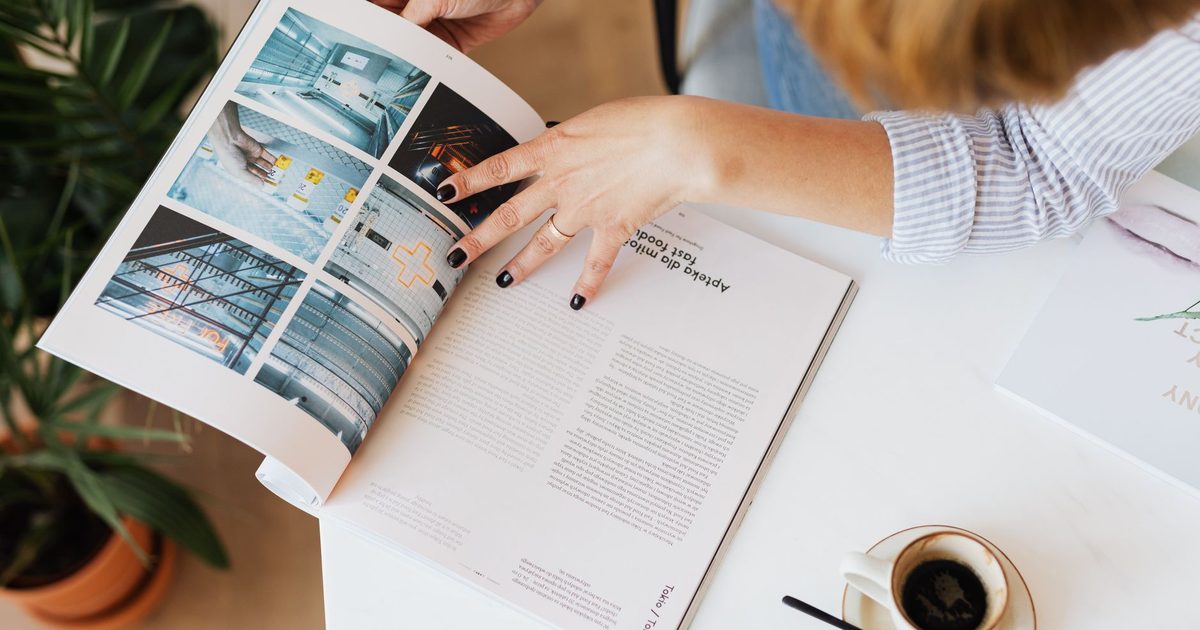 Hands turning the pages of a magazine on a desk beside a coffee cup