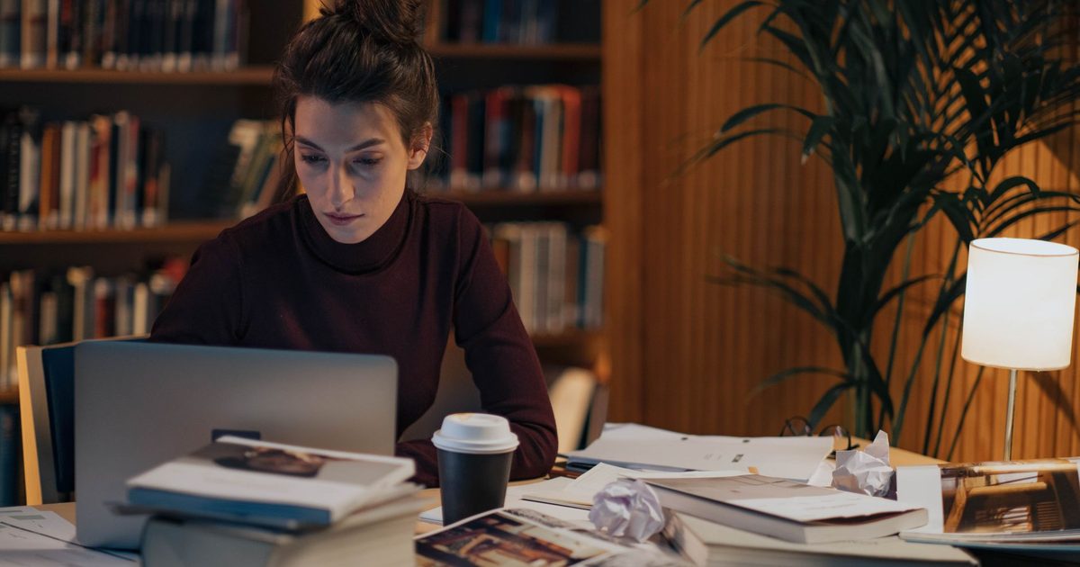 Woman working at a desk covered with open books and a laptop