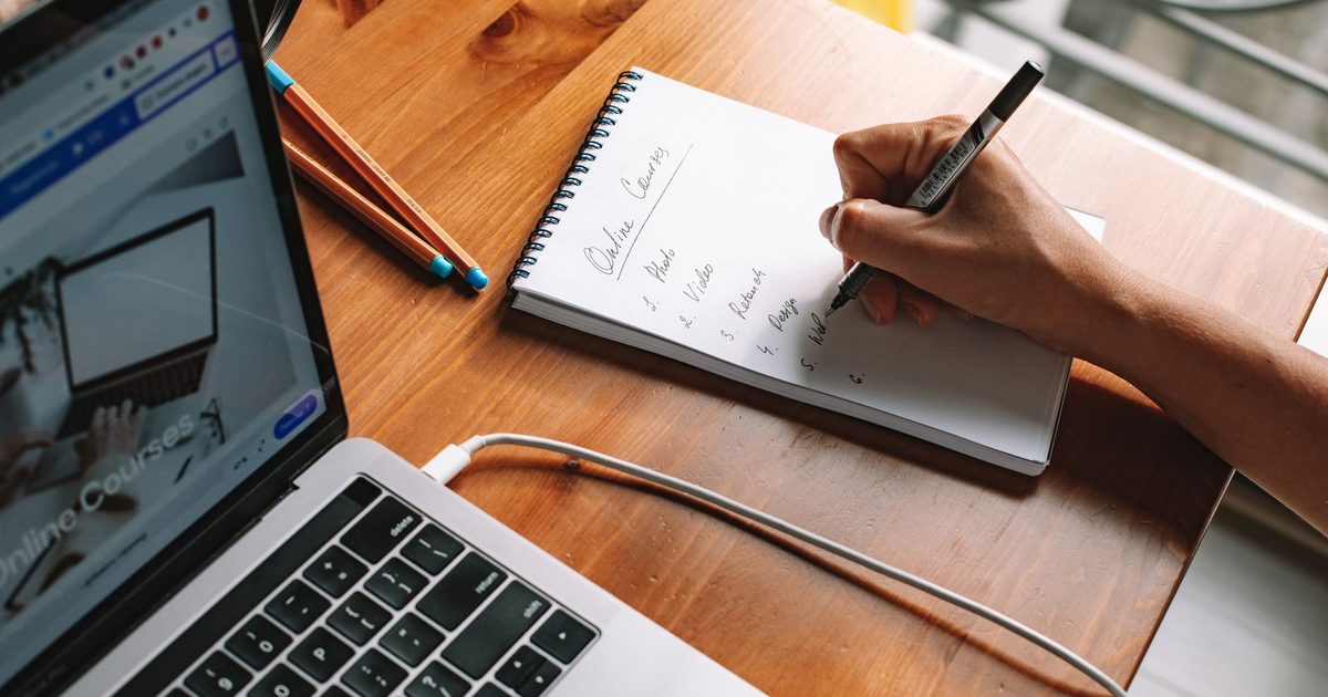 Hand writing notes in a notebook beside an open laptop